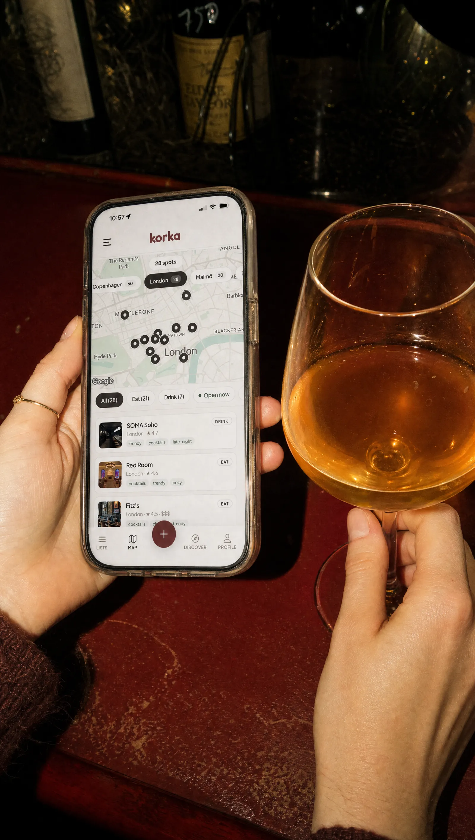 En hand som håller en telefon med Korkas karta, intill ett glas orange vin på en bardisk